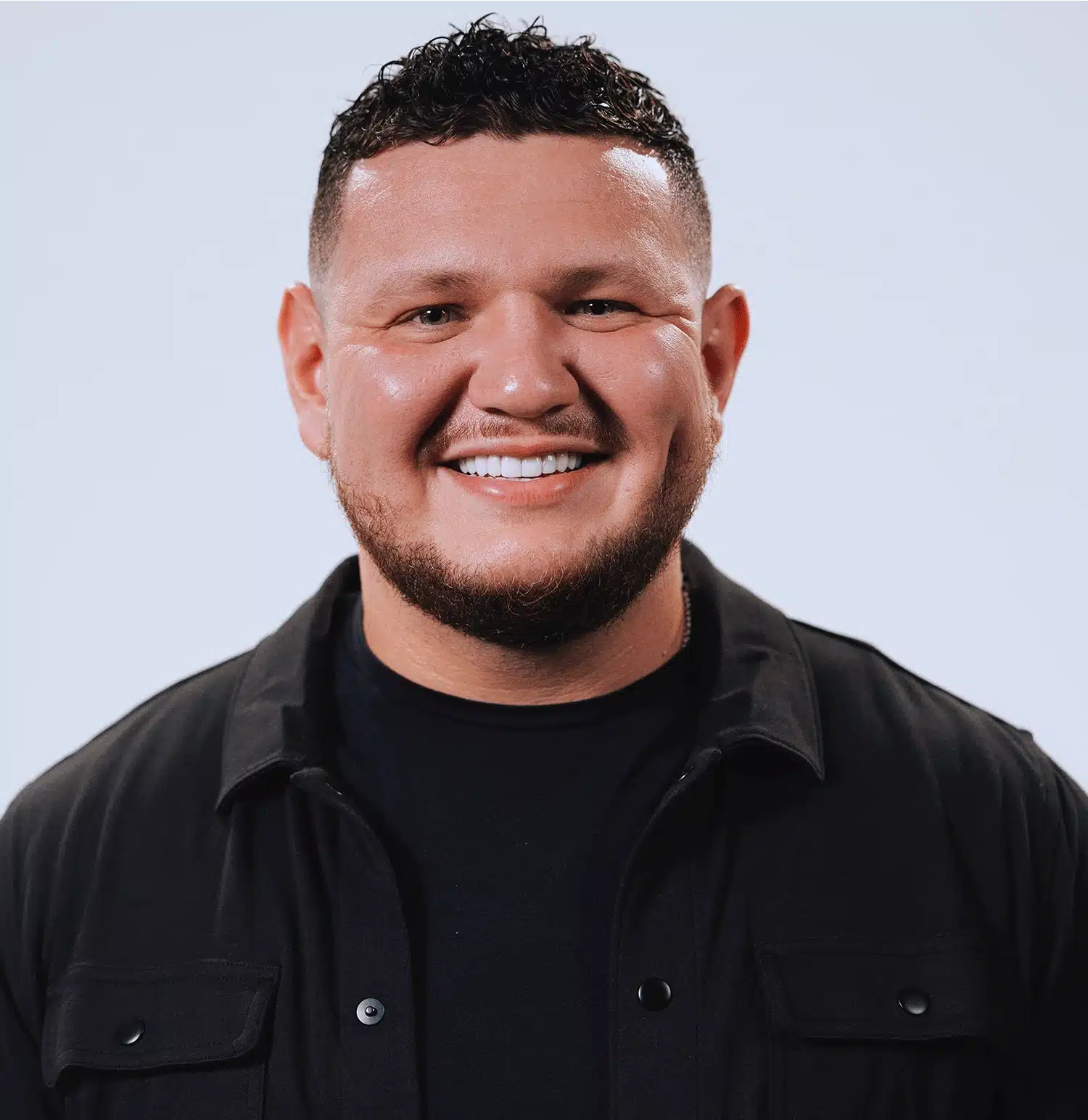 Smiling person with curly hair wearing a black jacket and t-shirt against a light background.