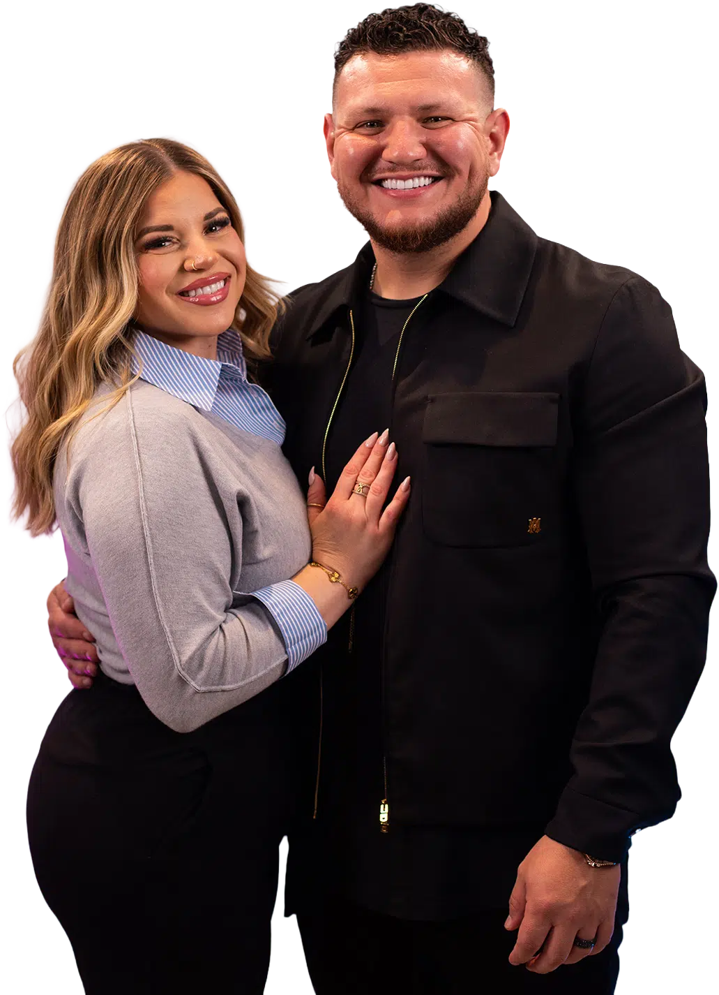 A couple stands together, smiling; she wears a light gray sweater, he a black jacket.
